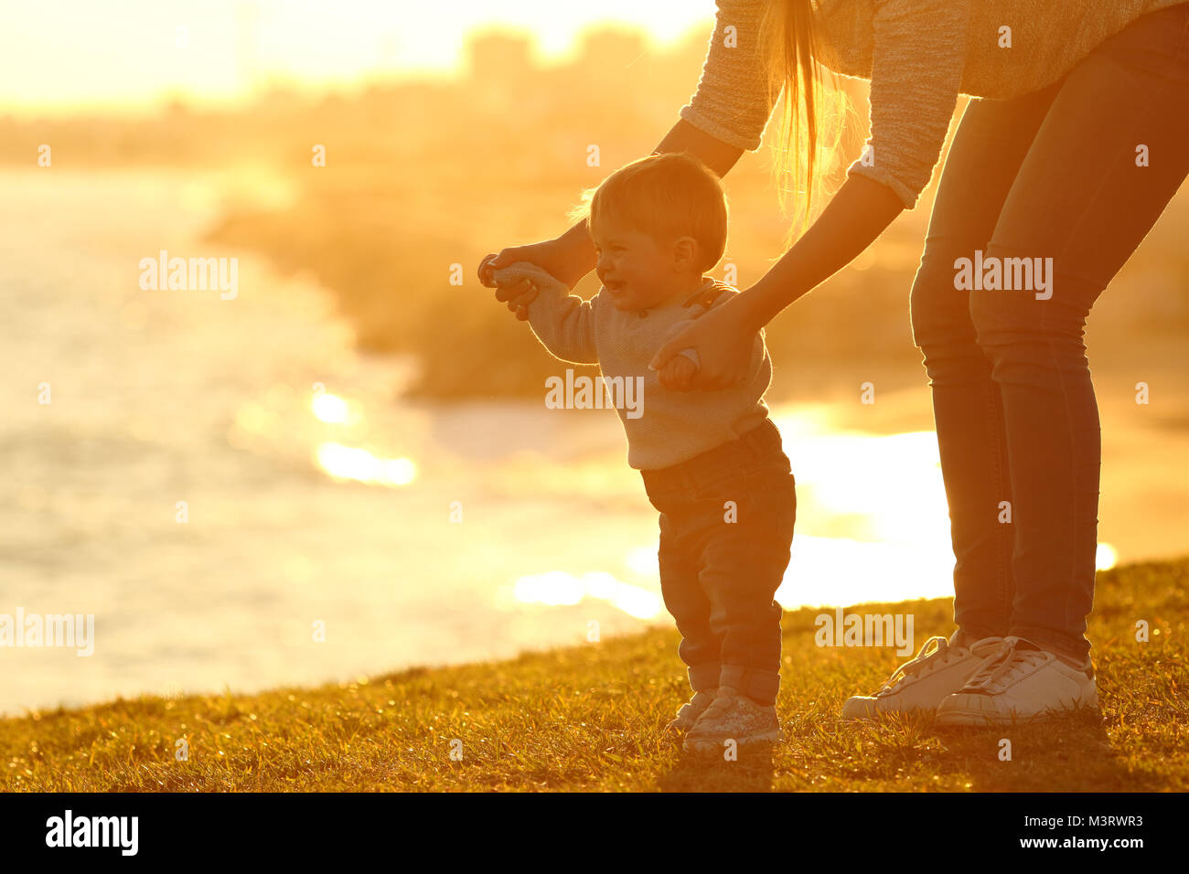 Vista laterale di un bambino ad imparare a camminare e madre aiutandolo sull'erba all'aperto al tramonto con una città in background Foto Stock