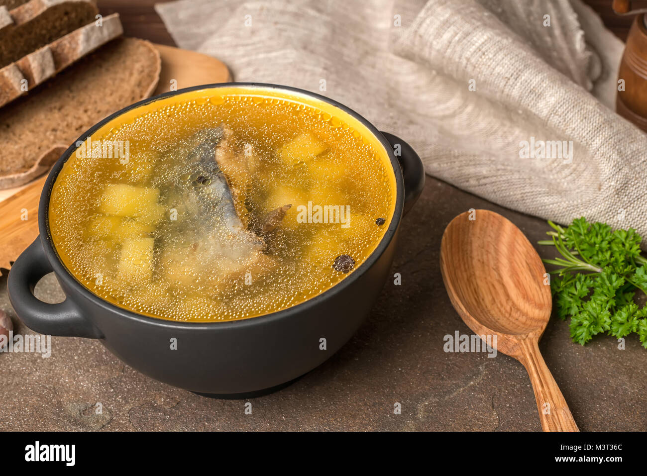 Trasparente la zuppa di pesce con lo storione, patate in piastra nera, decorato di aglio, prezzemolo in foglie, il cucchiaio di legno e tagliate il pane di segale, close up Foto Stock
