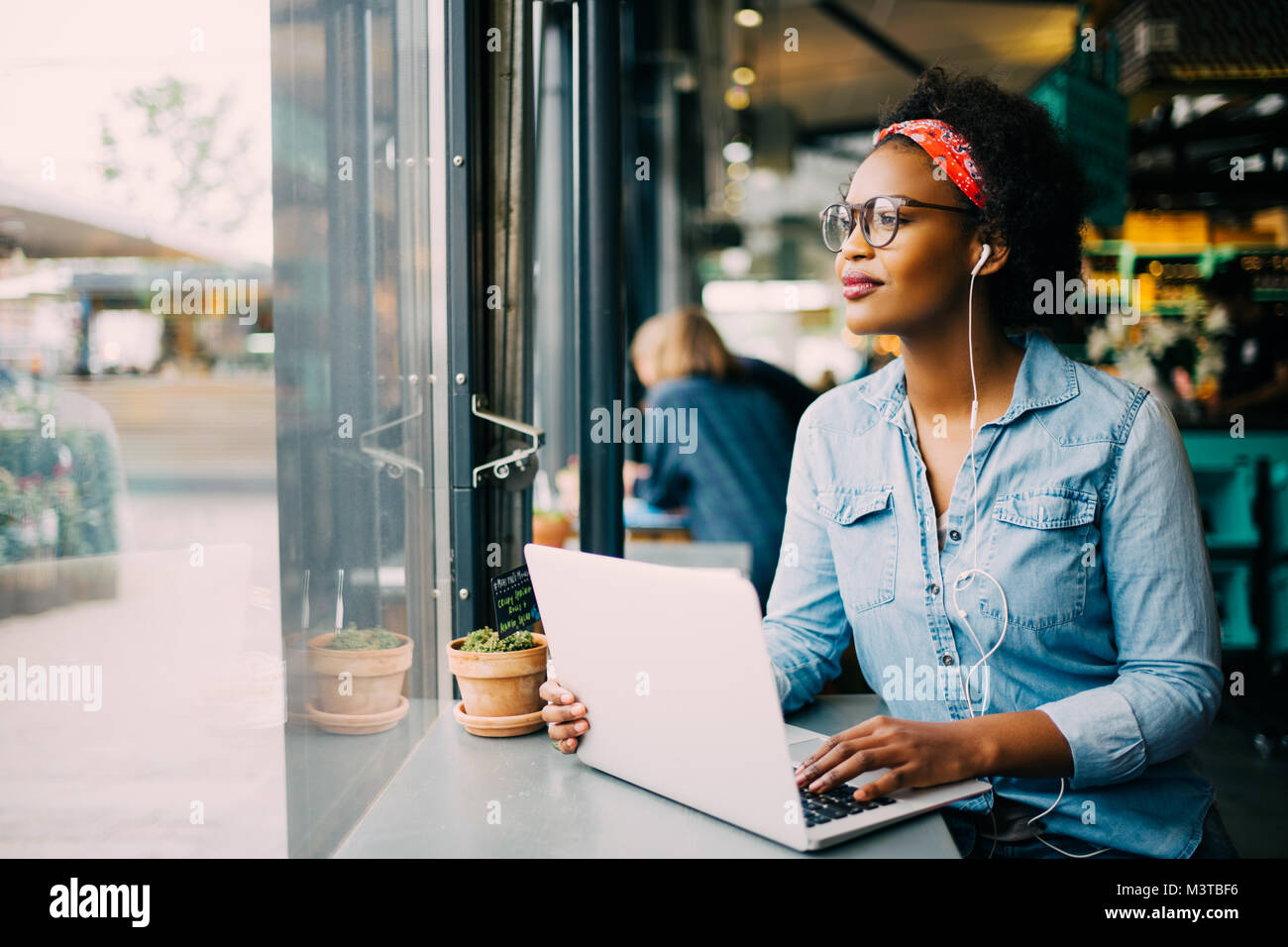 Sorridente giovane donna africana di guardare attraverso una finestra mentre è seduto da solo in cafe lavorando su un computer portatile e di indossare le cuffie Foto Stock