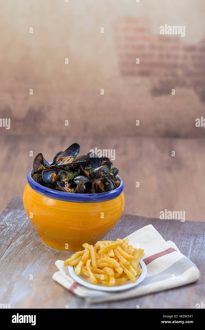 Blu francese cozza con le erbe in una vaschetta di colore giallo con patatine fritte di vetro su ramekin tovagliolo frutti di mare in una tavola di legno nella parete vintage background Foto Stock