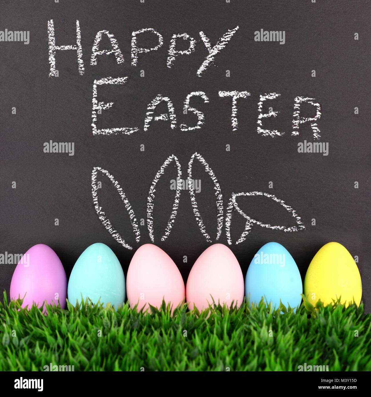 Felice Pasqua sulla lavagna con fila di uova di Pasqua in erba.Due con disegnati a mano coniglietto di Pasqua le orecchie. Foto Stock