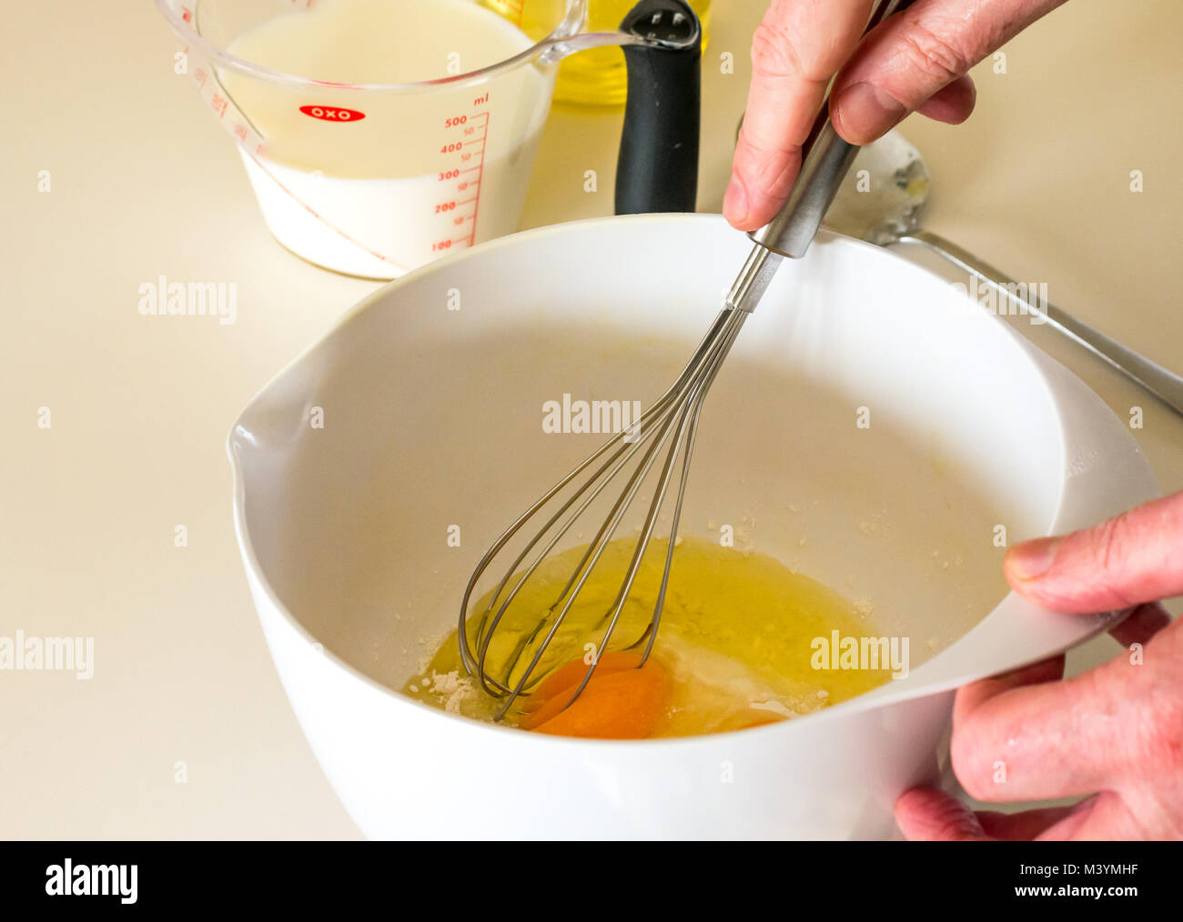 L'uomo battendo la miscela con una frusta per fare pastella di frittella dolce per il Martedì Grasso con farina, latte, uova e olio in una cucina domestica Foto Stock