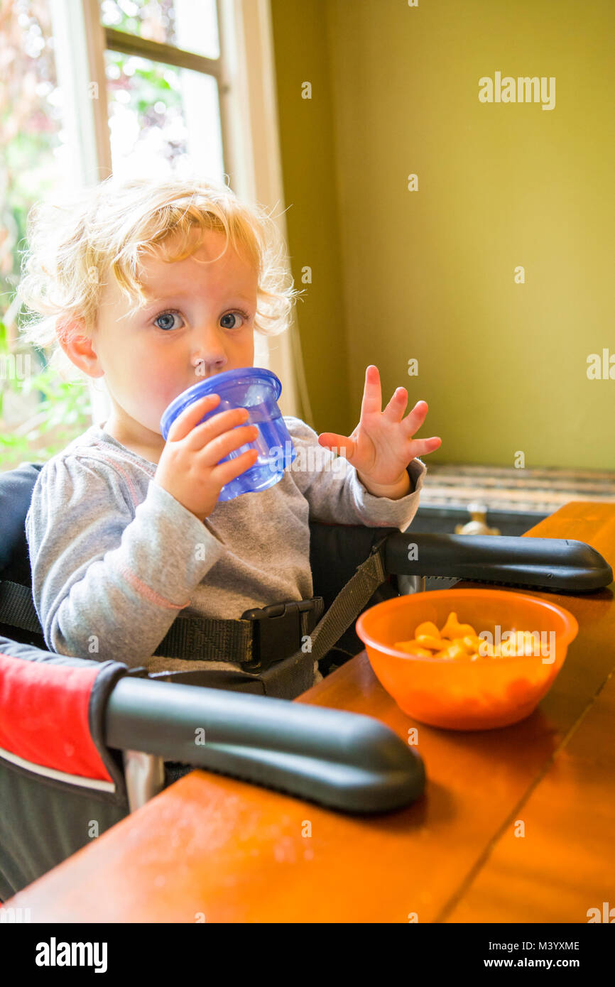 Un bambino ragazza seduta al tavolo con drink / sippy cup e una ciotola di cracker. Foto Stock