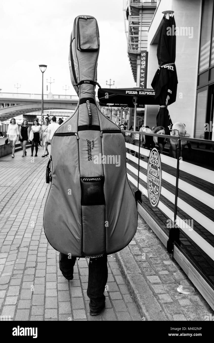 Londra in bianco e nero street photography: musicista a piedi nella strada che porta contrabbasso gig in borsa sul suo retro. Foto Stock