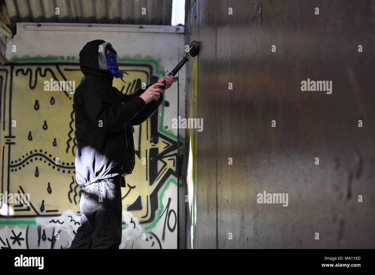 Un artista di graffiti con il suo volto coperto di verniciatura di un muro in un magazzino abbandonato con un rullo di vernice Foto Stock