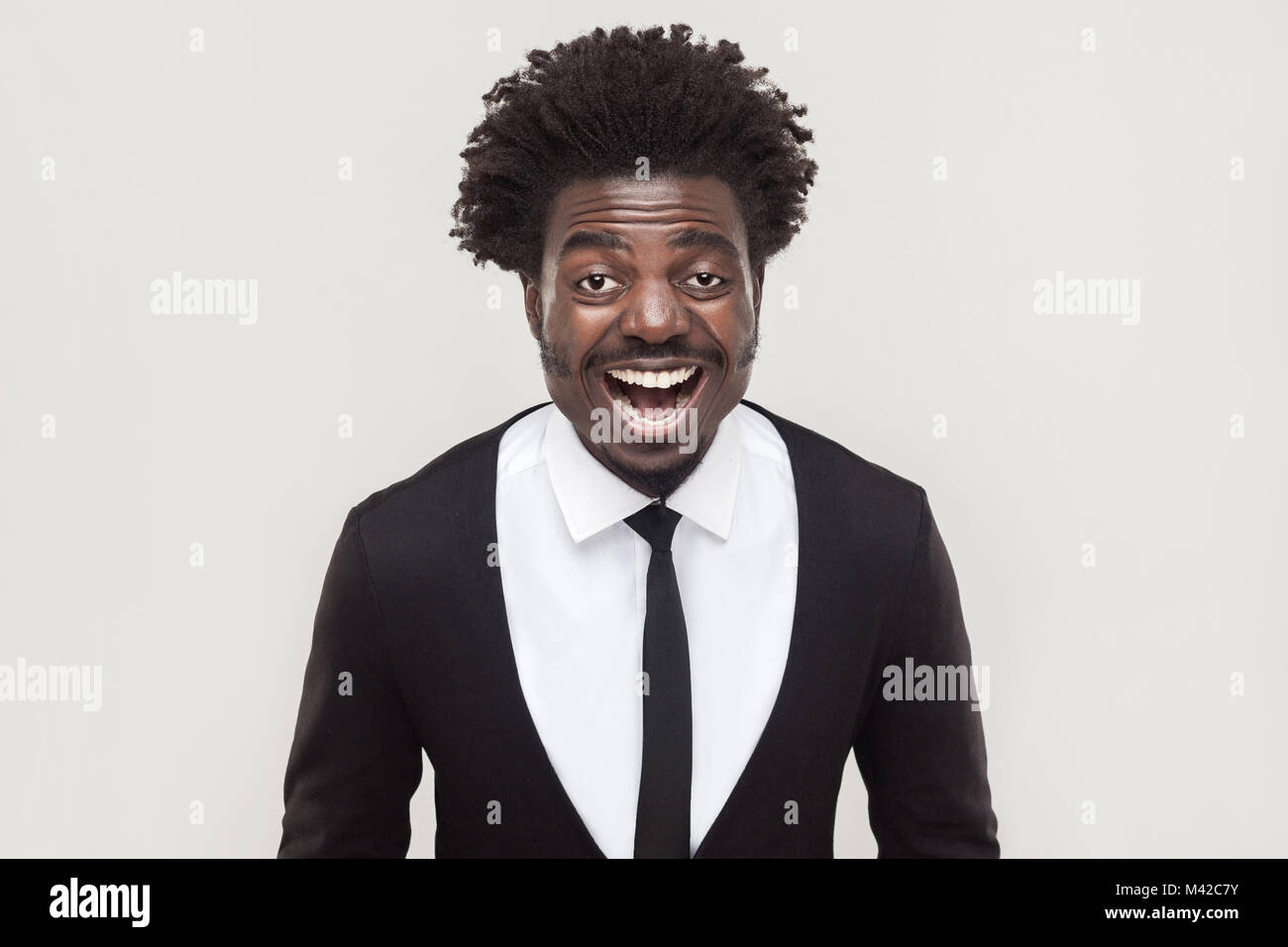 Ridendo afro imprenditore guardando la fotocamera e toothy sorridente. Studio shot, sfondo grigio Foto Stock