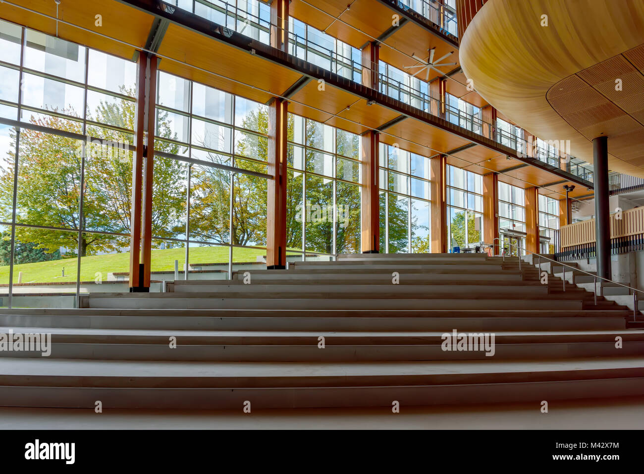 Una ampia scala con passi fino all'interno di un edificio moderno con colonne in legno, grandi finestre di vetro, erba verde, blu cielo e alberi Foto Stock