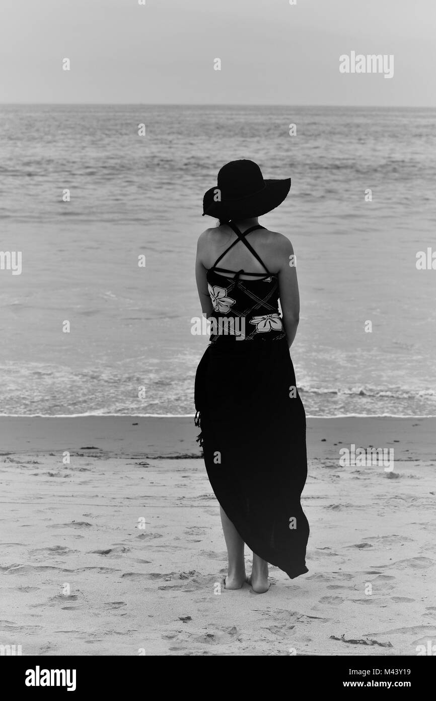 Giovani donne che indossano cappello permanente sulla spiaggia con la gonna al vento, bianco e nero, Stato Carpintaria Beach California Foto Stock