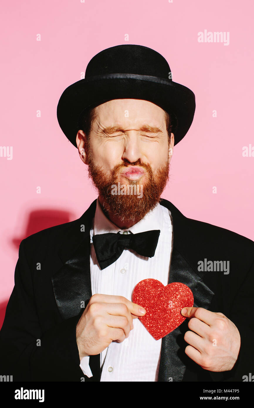 Funny guy in tux e il top hat busbana francese un bacio e tenendo un cuore rosso Foto Stock