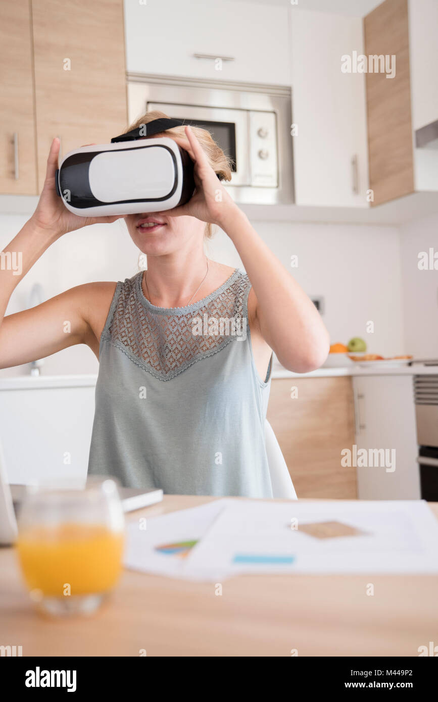 Giovane donna guardando attraverso la realtà virtuale auricolare a tavolo da cucina Foto Stock