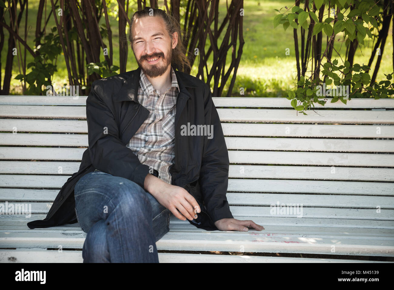 Giovani barbuto uomo felice sul banco di bianco in estate park, ritratto all'aperto Foto Stock