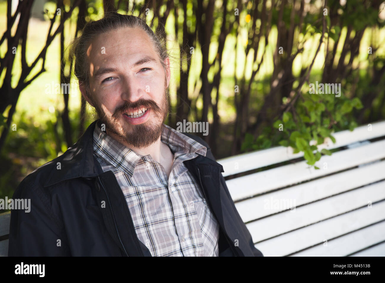 Giovani barbuto uomo sorridente sul banco di bianco in estate park, ritratto all'aperto Foto Stock