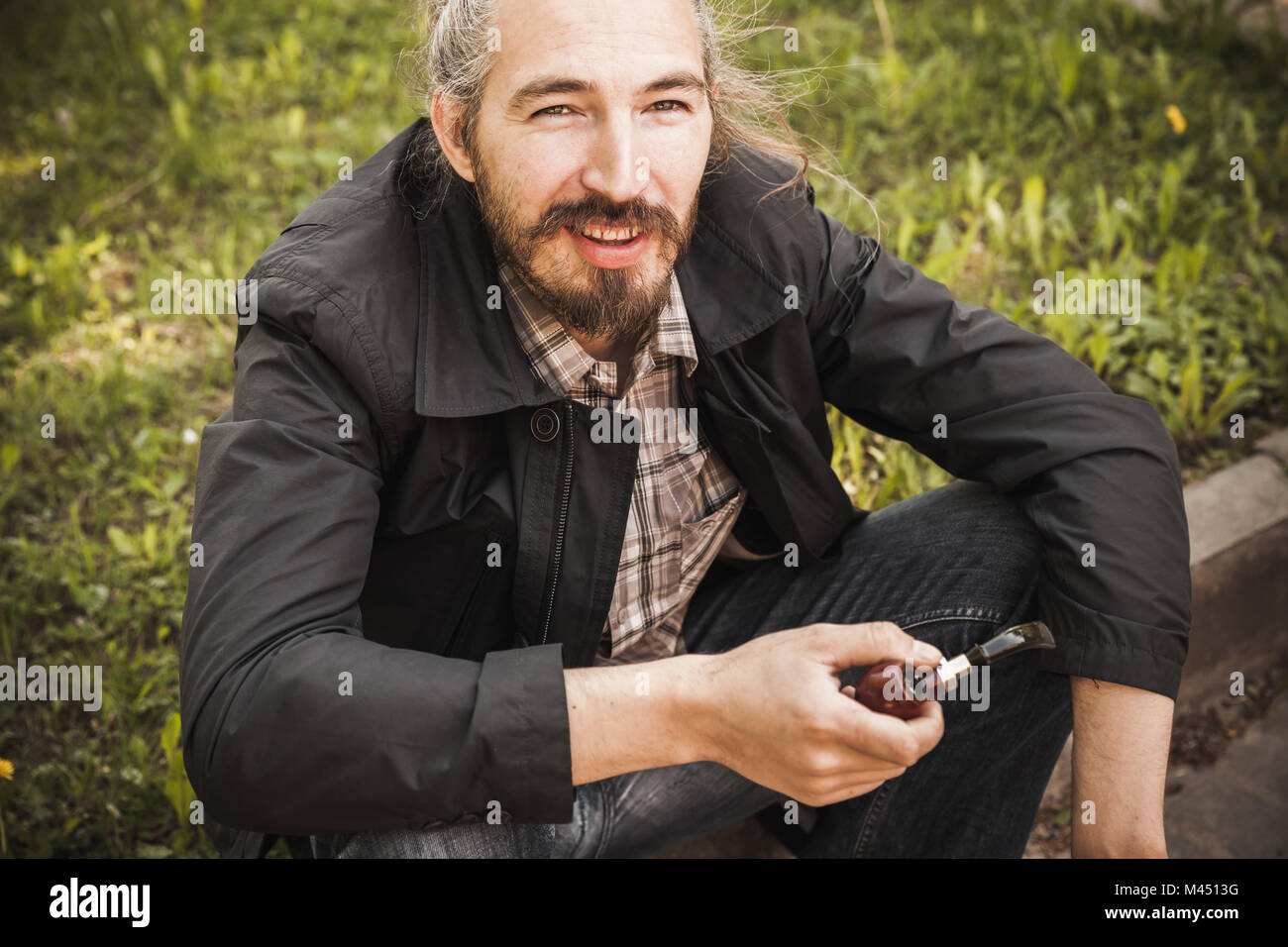 Giovani sorridente uomo barbuto tubazione di fumare in estate park, ritratto all'aperto Foto Stock