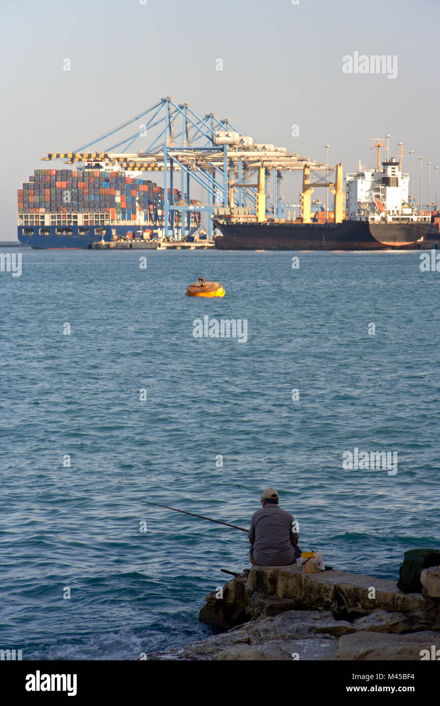 Un uomo pesca con vista verso il grande contenitore morsetti della Freeport a Birzebugga, Malta. Foto Stock