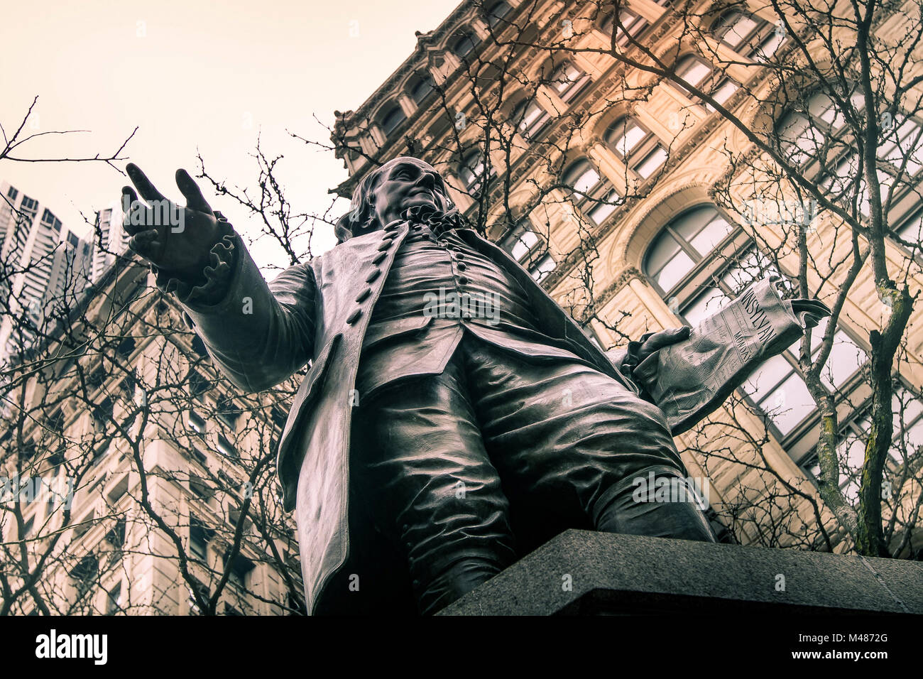 Vista verso l'alto di una statua di Benjamin Franklin con rami di alberi ed edifici sullo sfondo. Foto Stock