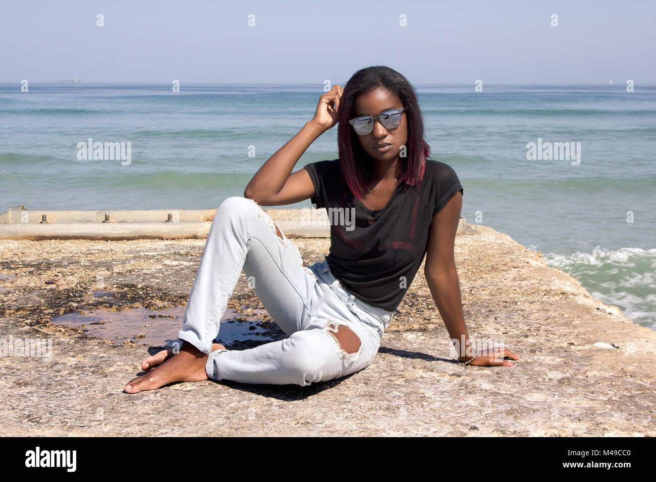 Elegante donna etnica in jeans e maglietta e occhiali da sole seduto su di un molo di cemento dall'oceano guardando la telecamera Foto Stock