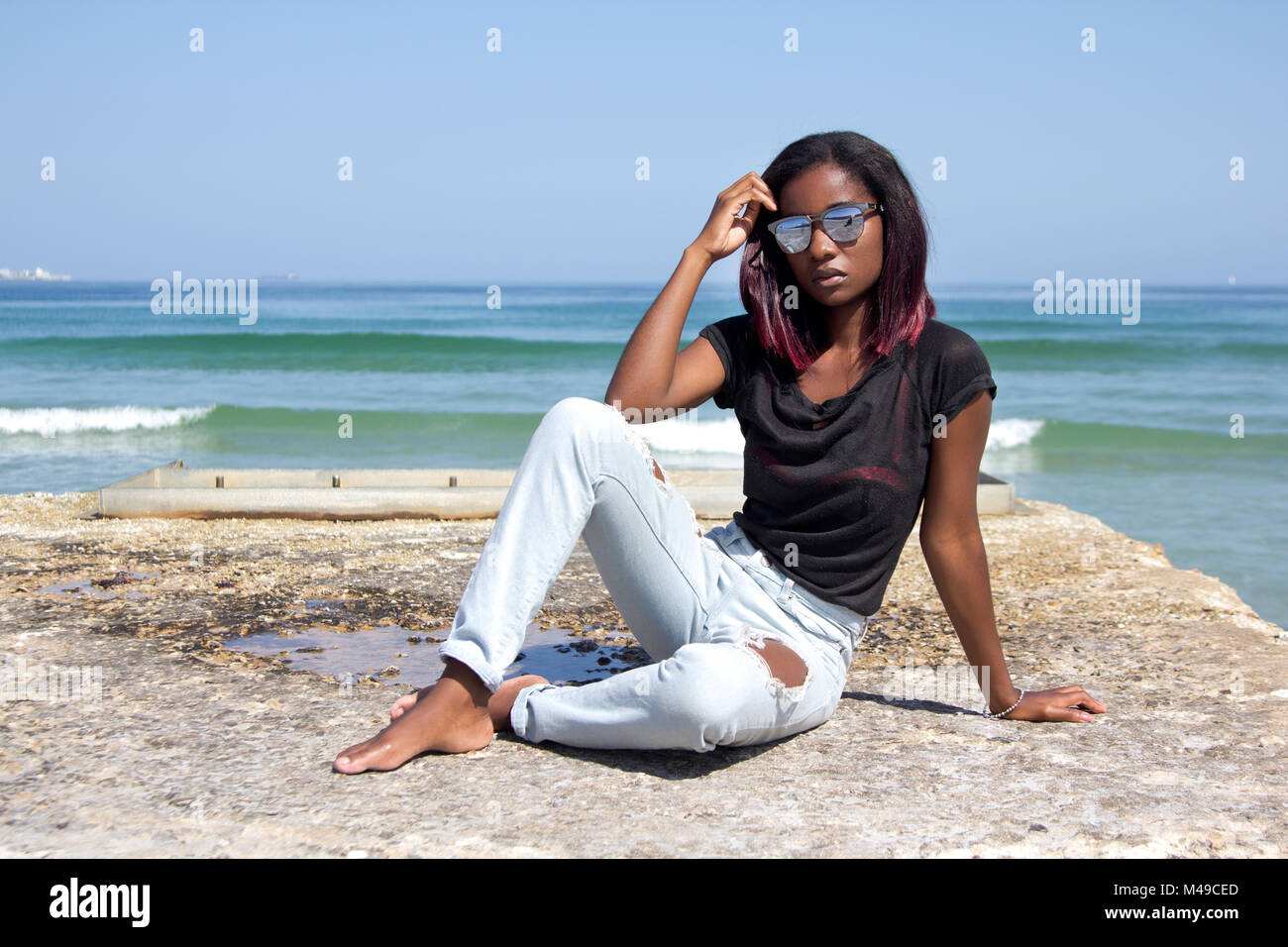 Elegante donna etnica in jeans,camicia e occhiali da sole seduto su di un molo di cemento dall'oceano guardando la telecamera con copyspace Foto Stock