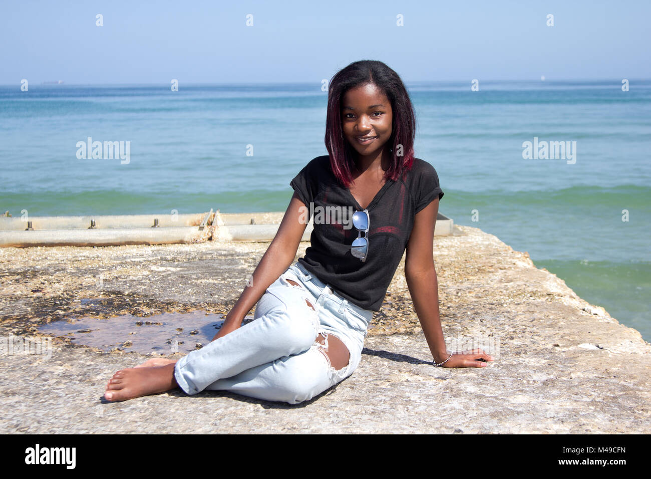 Un felice ragazza africana in jeans e una maglietta e occhiali da sole seduto su di un molo di cemento dall'oceano Foto Stock