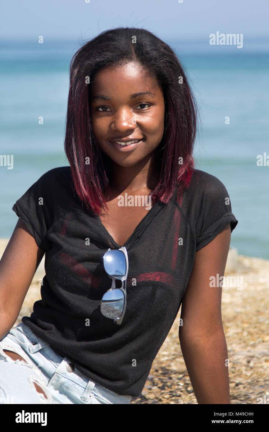 Ritratto di un felice ragazza africana in jeans e una maglietta e occhiali da sole seduto su di un molo di cemento dall'oceano Foto Stock