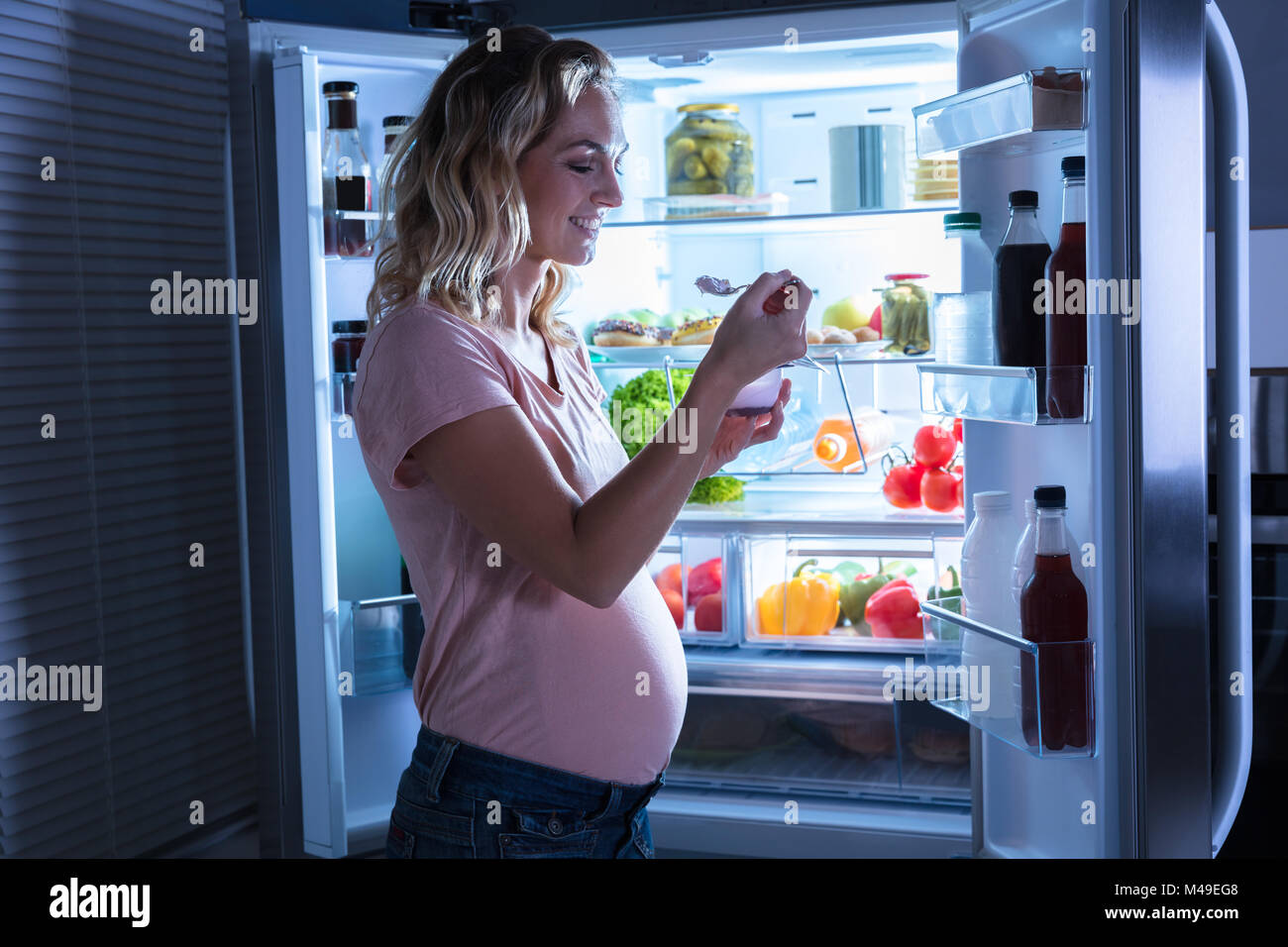 Giovane donna incinta a mangiare il gelato nella parte anteriore del frigorifero aperto Foto Stock