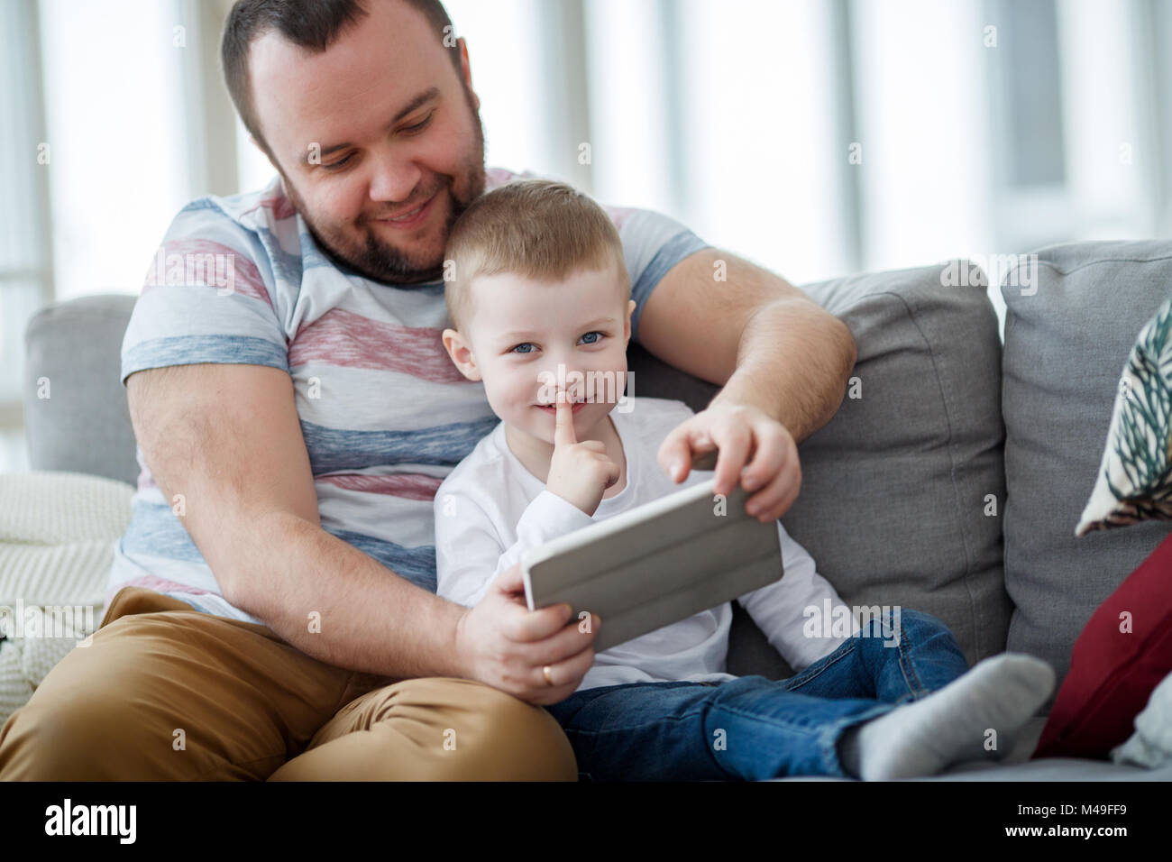 Foto di felice papà con suo figlio con la compressa seduto sul divano grigio Foto Stock