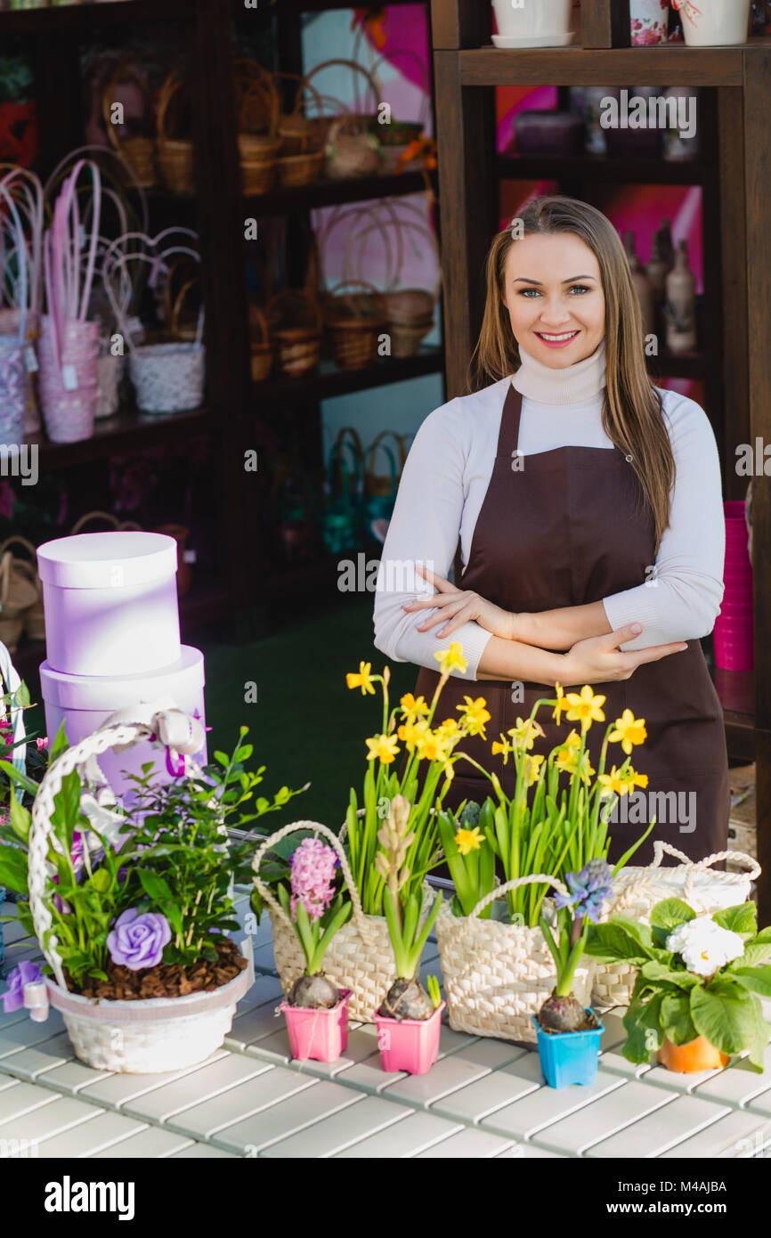 Il venditore di un negozio di fiori, in piedi accanto al tavolo con fiori, attraversato le sue mani e sorridente, guardando la telecamera Foto Stock