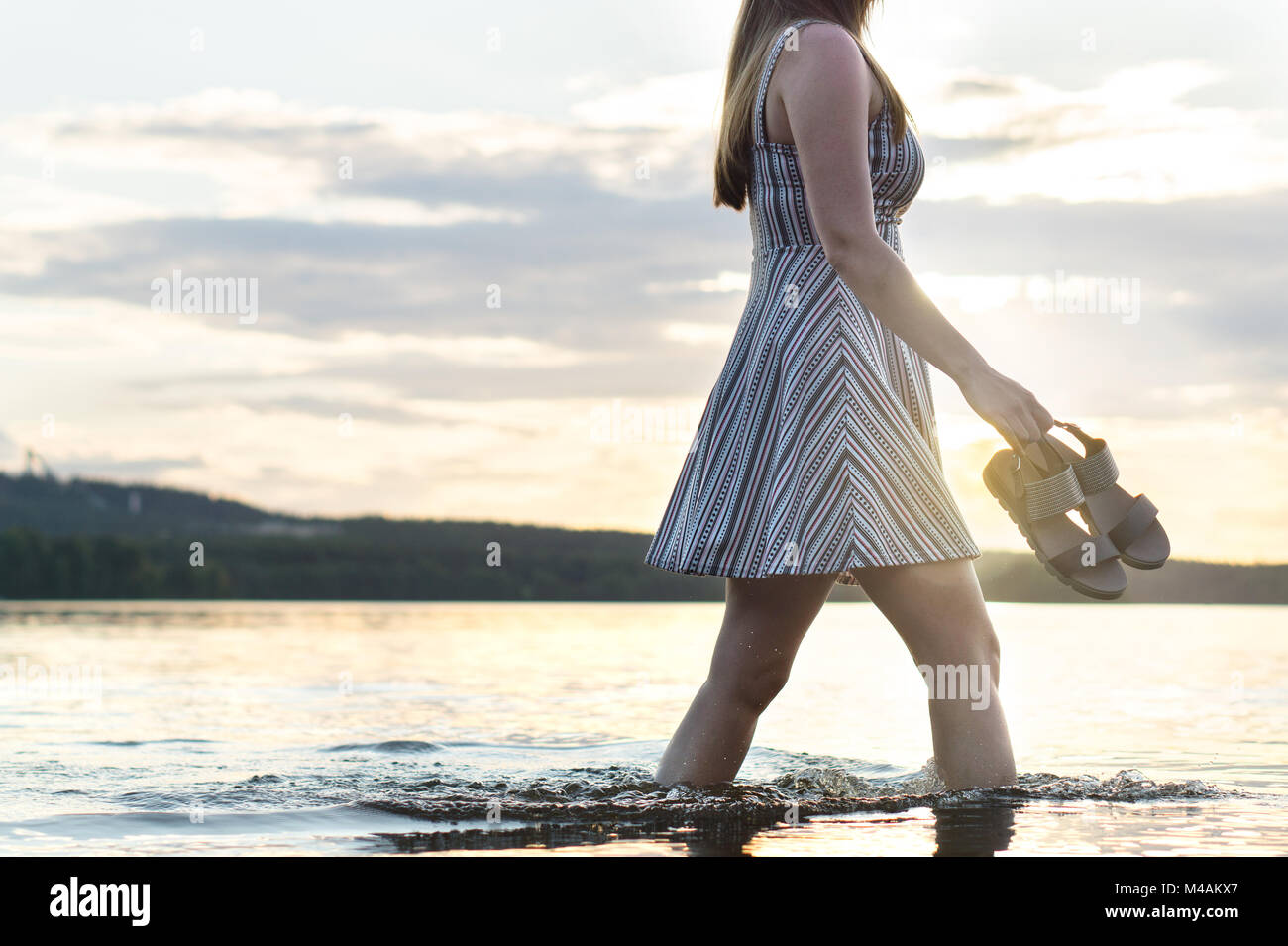 Giovane donna attraente a piedi in acqua di lago al tramonto. Spensierato e felice di stile di vita. Azienda sandali in mano. La libertà e la felicità del concetto. Foto Stock
