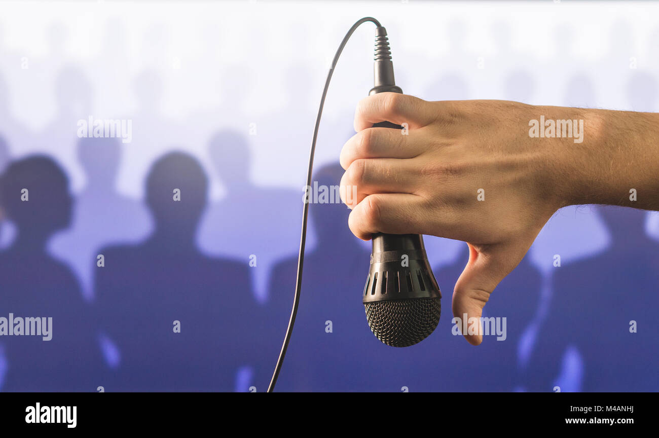 Mano che tiene il microfono e mostrando il pollice verso il basso davanti a una folla di persone di silhouette. Impossibile parlare in pubblico e dare la parola concetto. Foto Stock