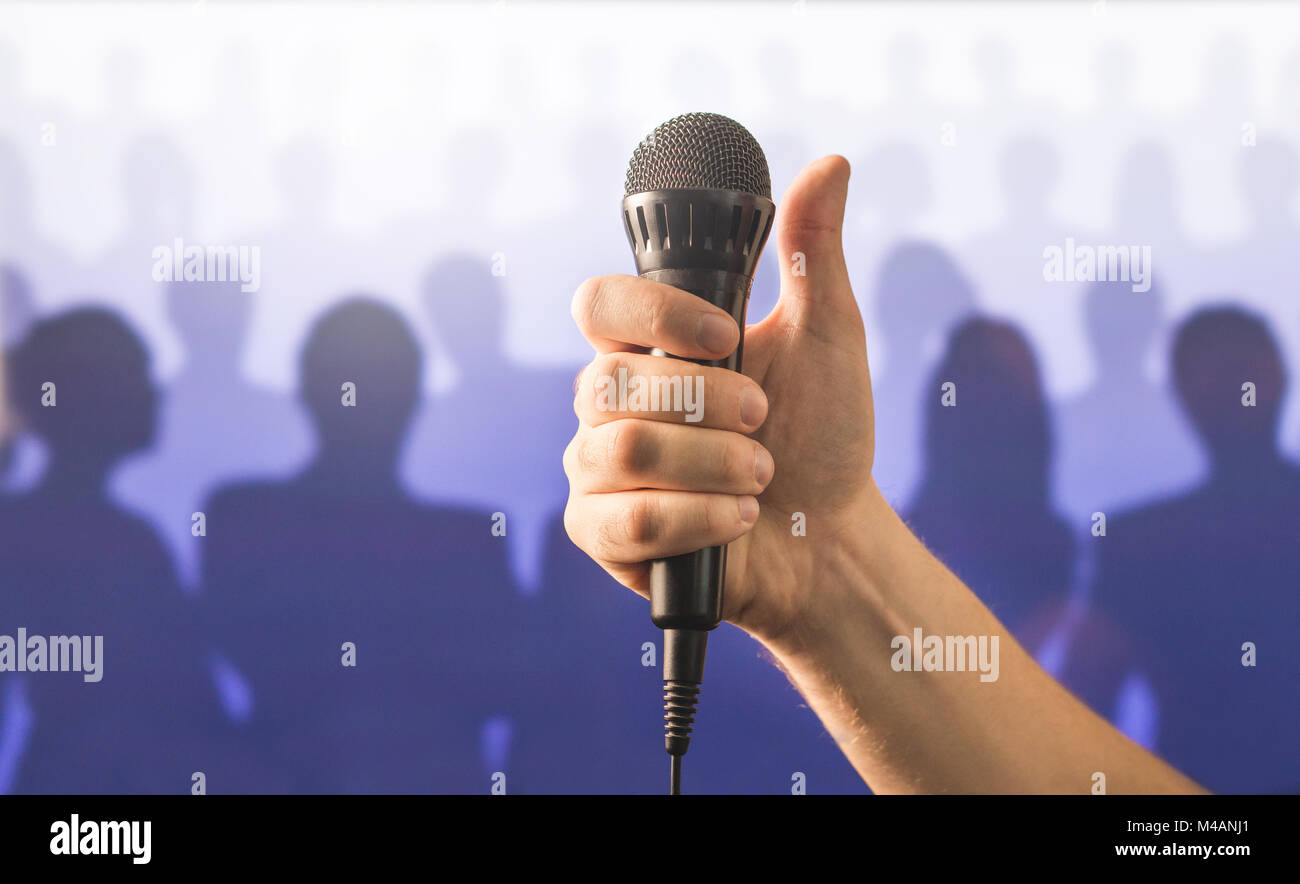 Mano che tiene il microfono e che mostra i pollici fino davanti a una folla di persone di silhouette. Parlare in pubblico e dare la parola concetto. Canti bene vivere. Foto Stock