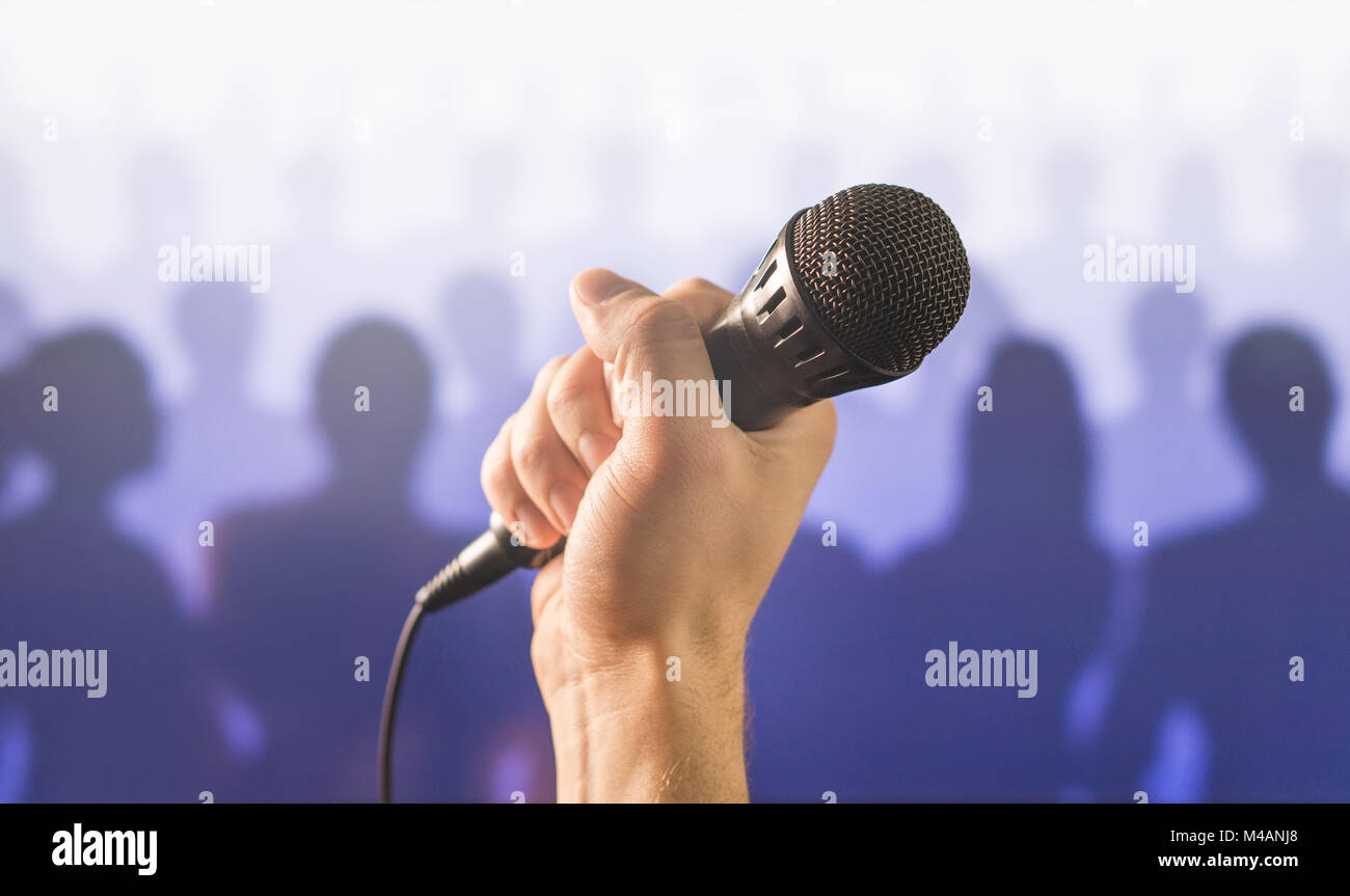 Parlare in pubblico e dare la parola concetto. Mano che tiene il microfono davanti a un pubblico di silhouette e la folla di gente. Il canto a microfono karaoke. Foto Stock