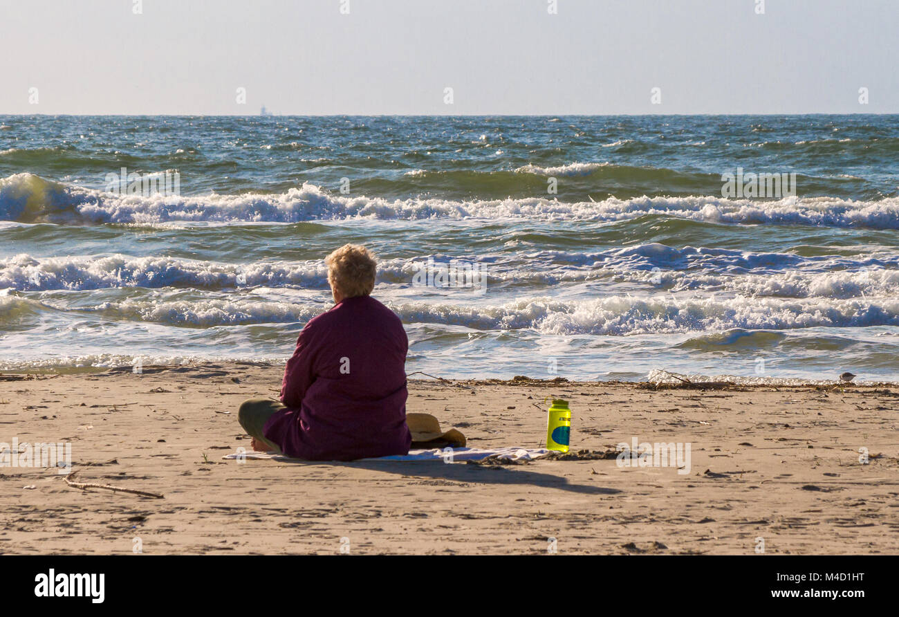 Un Americano senior citizen donna siede sulla spiaggia e guarda all'oceano nelle prime ore del mattino la luce del sole. Foto Stock