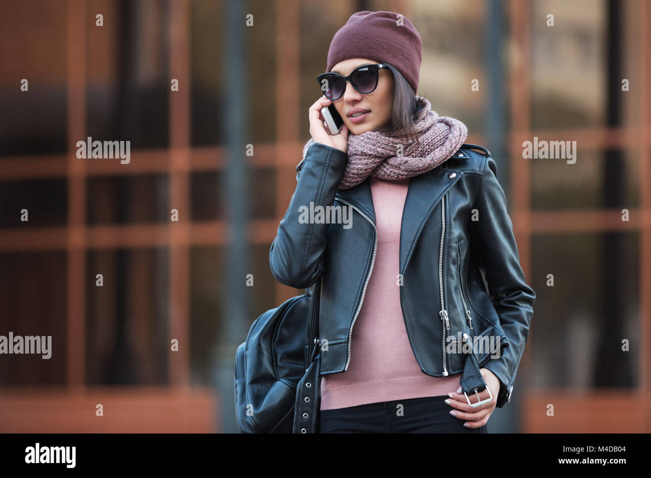 Moda giovane donna in nero giacca di pelle utilizzando il cellulare in una strada di città elegante modello femminile di indossare occhiali da sole beanie e sciarpa per esterno Foto Stock