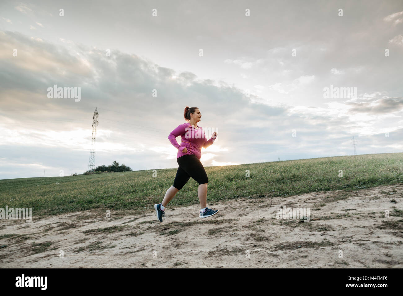 Basso angolo di visione di un paffuto pareggiatore femmina jogging fino alla cima di una collina al tramonto. Foto Stock