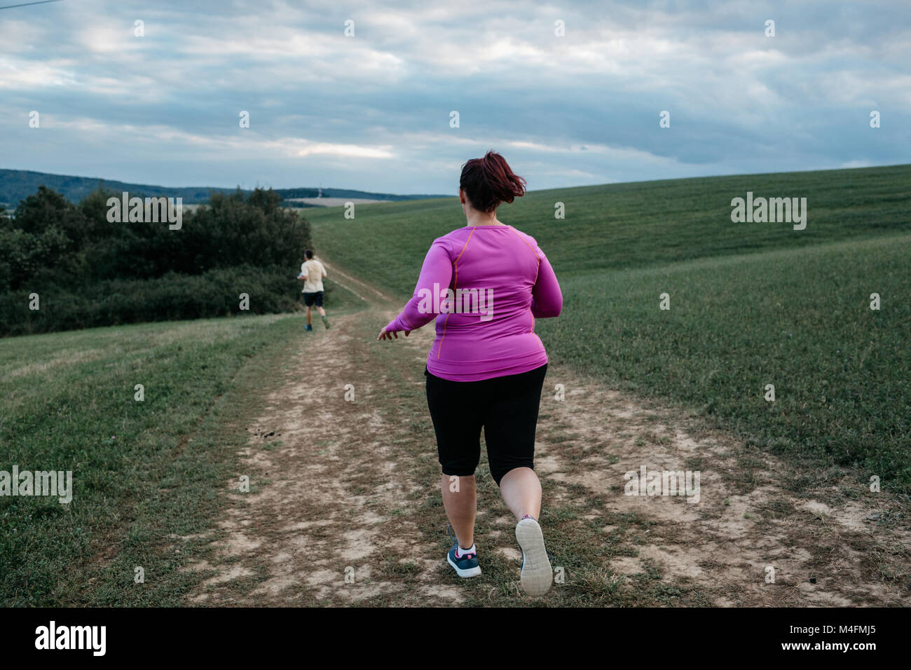 Vista posteriore di una donna sovrappeso nell'abbigliamento sportivo in esecuzione dopo un uomo lungo un sentiero del paese. Foto Stock