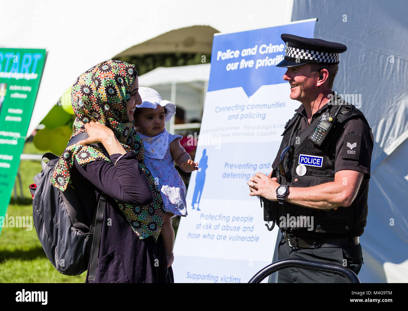 Un funzionario di polizia e una donna musulmana con un bambino piccolo a stare di fronte ad un crimine prevenzione banner pur impegnandosi in una conversazione amichevole. Foto Stock