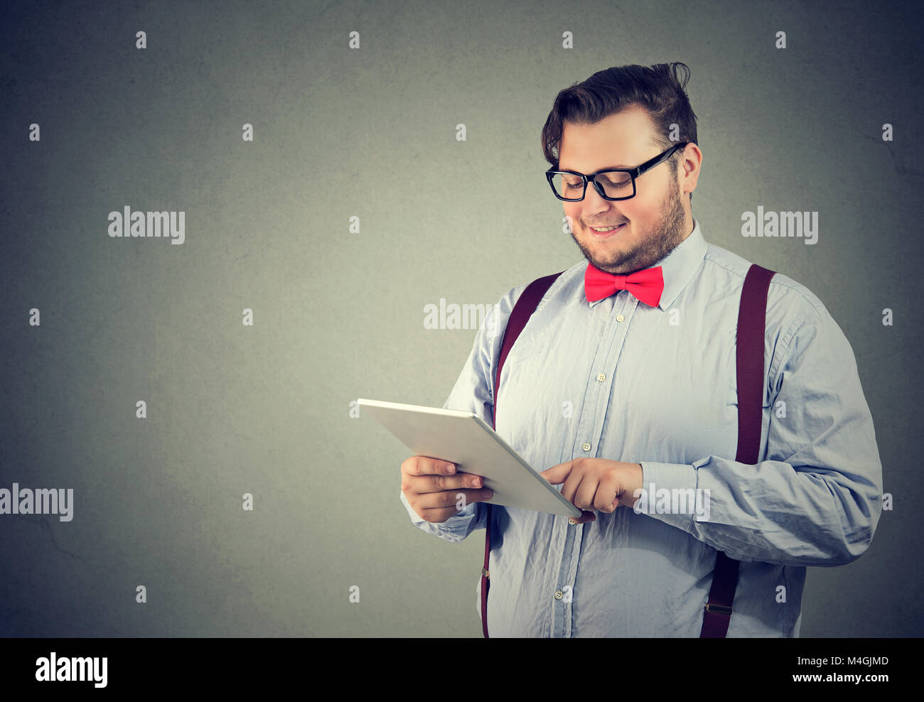 Giovane uomo cheeky in occhiali cercando felice durante la maschiatura tablet in posa su uno sfondo grigio. Foto Stock