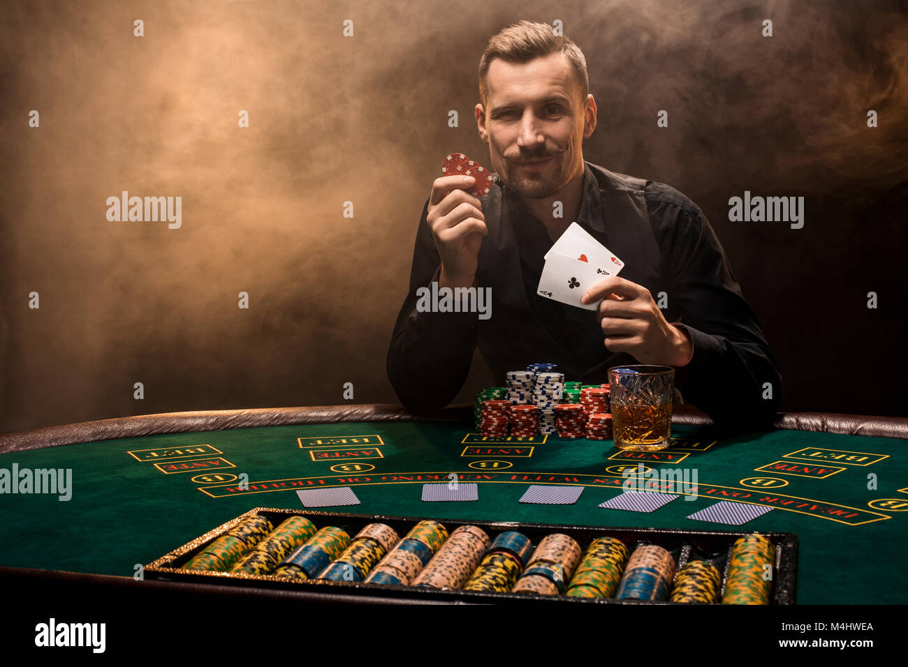 Bello il giocatore di poker con due assi nelle sue mani e patatine seduti al tavolo del poker in una stanza buia piena di fumo di sigaretta. Schede, trucioli, whisky, sigari Foto Stock