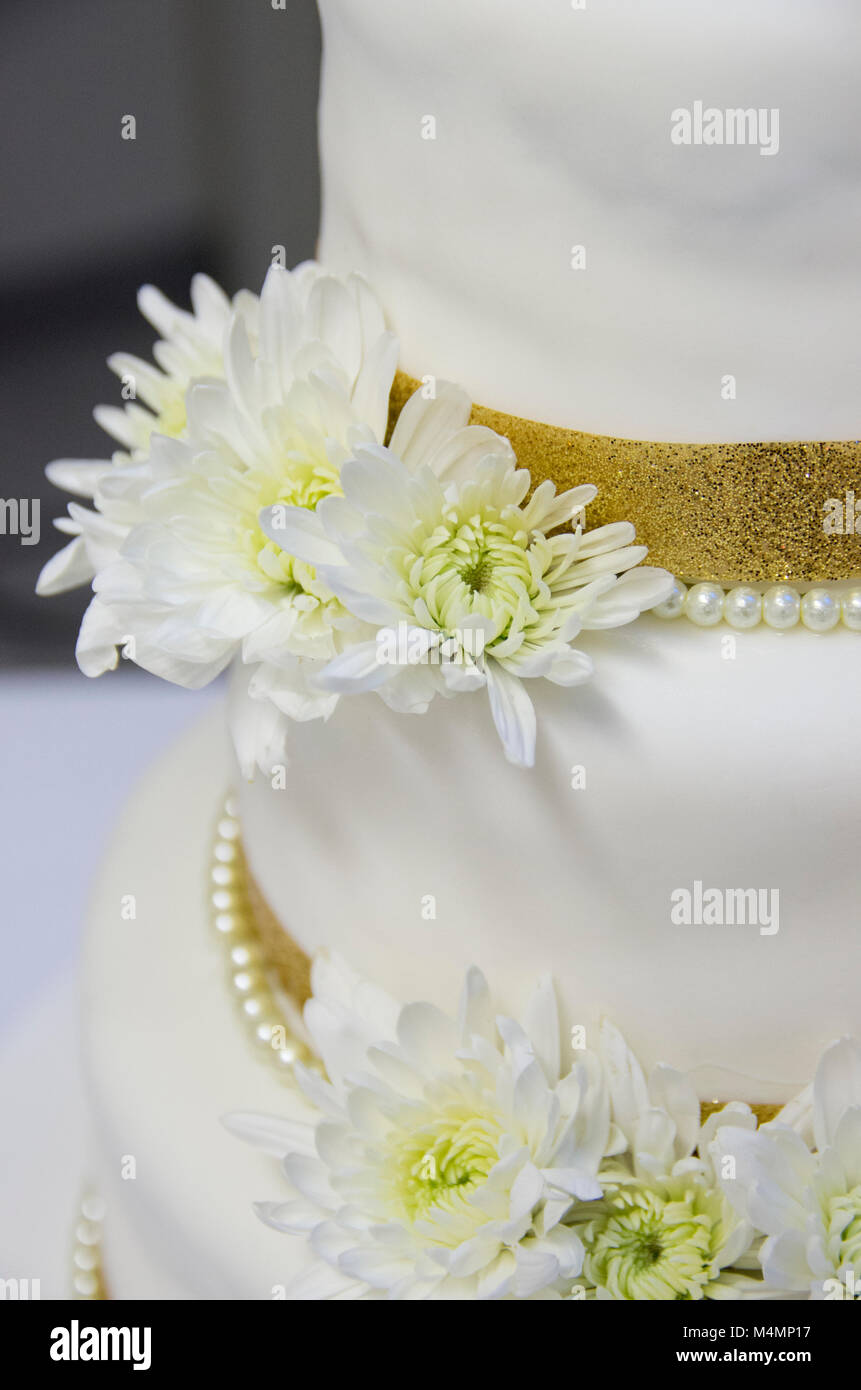 Porzione di satinato bianco torta di nozze con fresca crisantemo bianco fiori, anche un oro sfavillavano band e stringa di perle bianche intorno a ciascun livello. Foto Stock