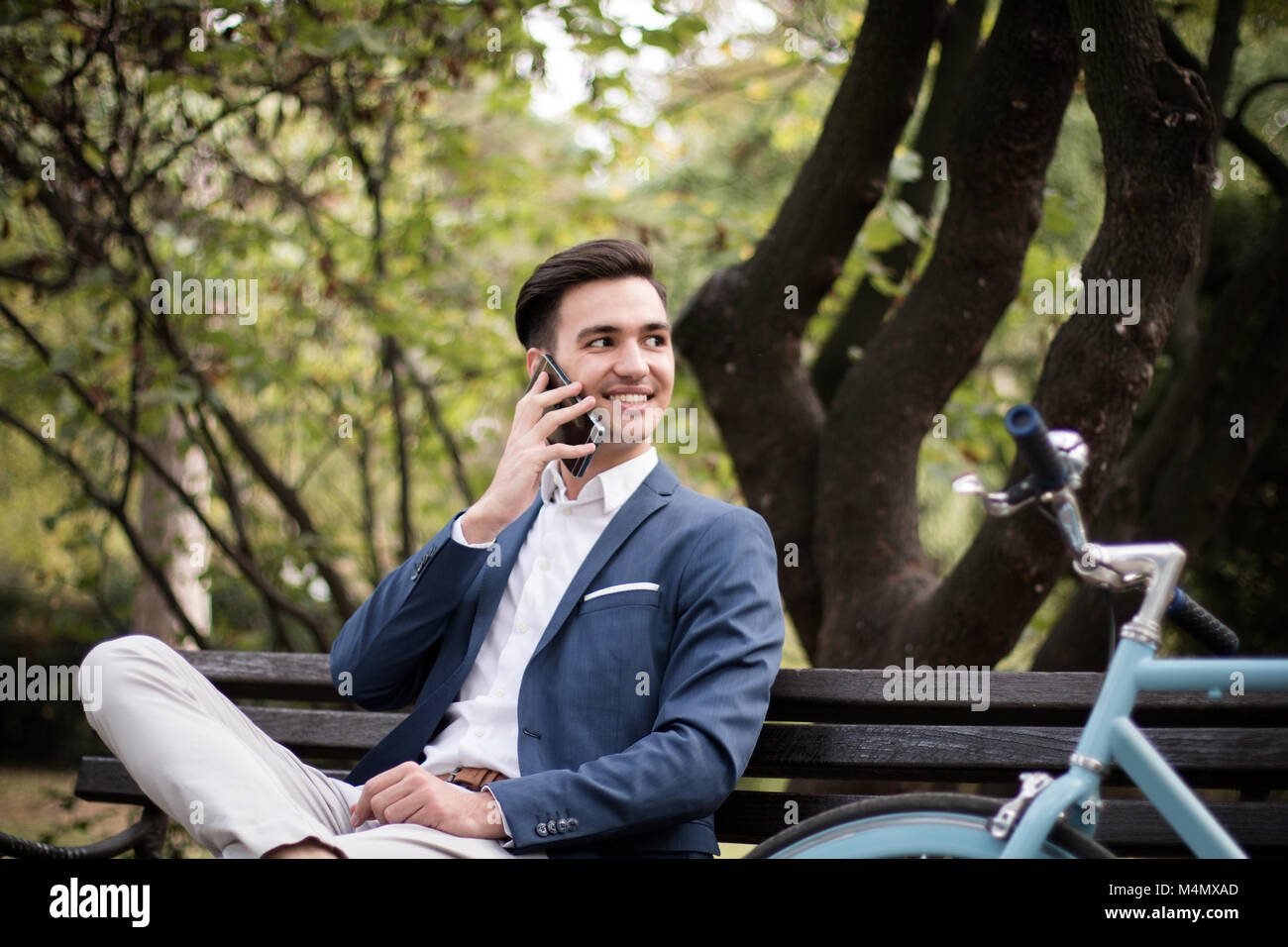 Giovane imprenditore parlando sullo smartphone all'aperto in un parco, seduta su una panchina con la sua bicicletta accanto a lui. Foto Stock