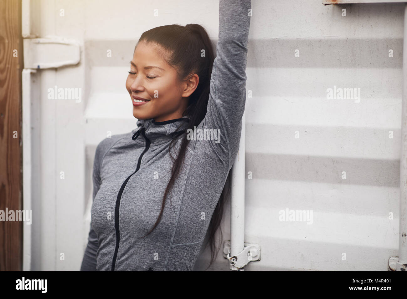Sorridente giovane donna asiatica in sportswear appoggiata contro un muro esterno con gli occhi chiusi e il braccio in aria prima di esercizio Foto Stock