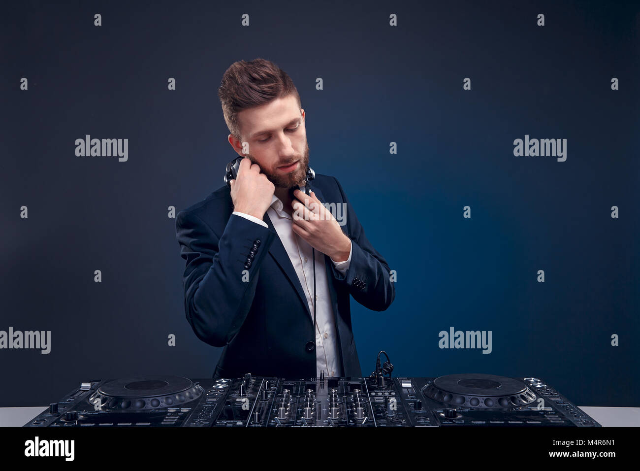 Closeup ritratto di certi DJ con elegante stile di capelli e cuffie sul collo di musica di miscelazione in mescolatore mentre in piedi isolato su di colore scuro blu, sfondo ciano Foto Stock