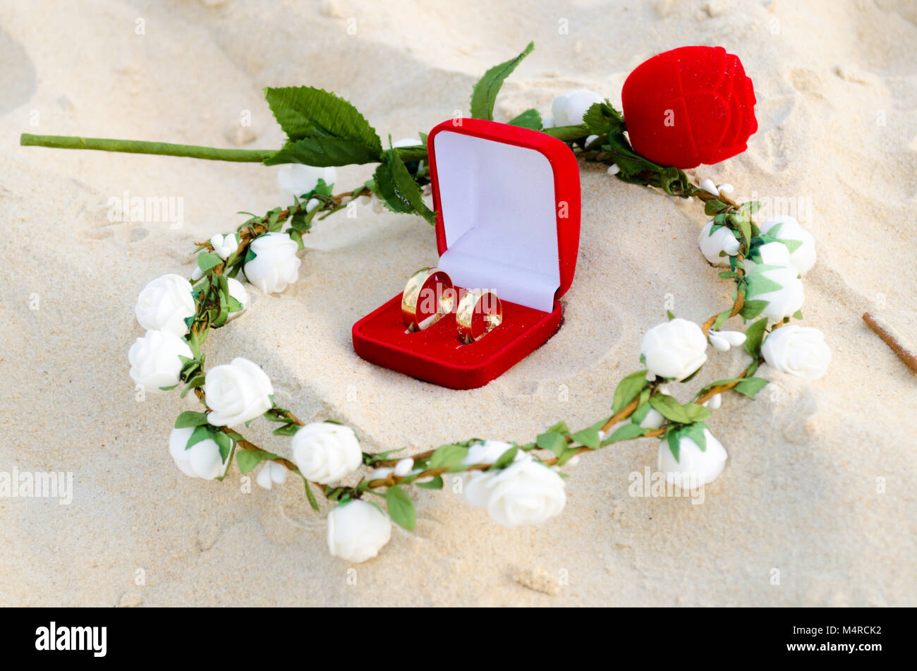 Casella rossa con gli anelli di nozze al centro di una ghirlanda di fiori bianchi sulla sabbia e una piccola rosa in background Foto Stock