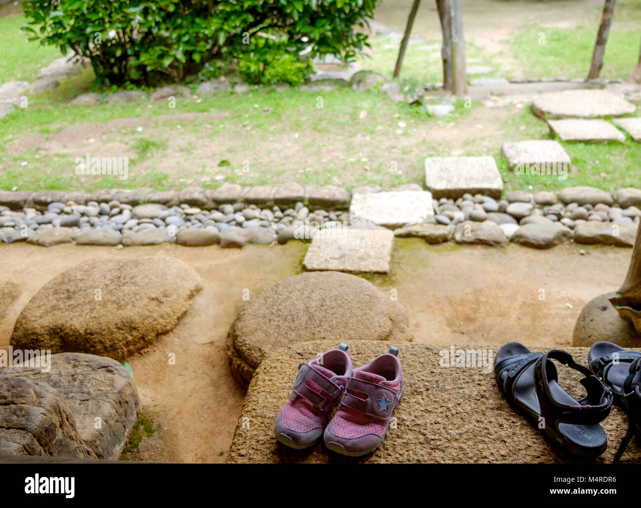 Rosa calzature per bambini sul gradino di pietra. Sulla destra sono nero sandali. Sullo sfondo a sinistra sono alberi. Sulla destra la parte inferiore di una struttura in stile rustico. Foto Stock