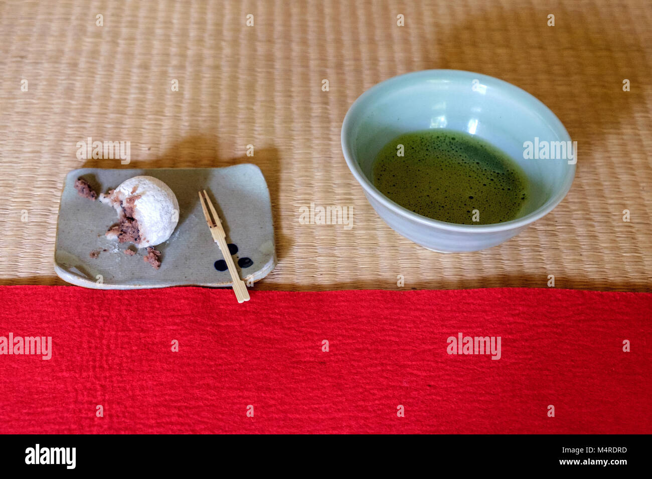 Tradizionale dolce giapponese su una piastra con una forcella di legno sulla sinistra & verde Matcha tè in una tazza di tè sulla destra. Collocato su di bambù e tappeti rossi. Foto Stock
