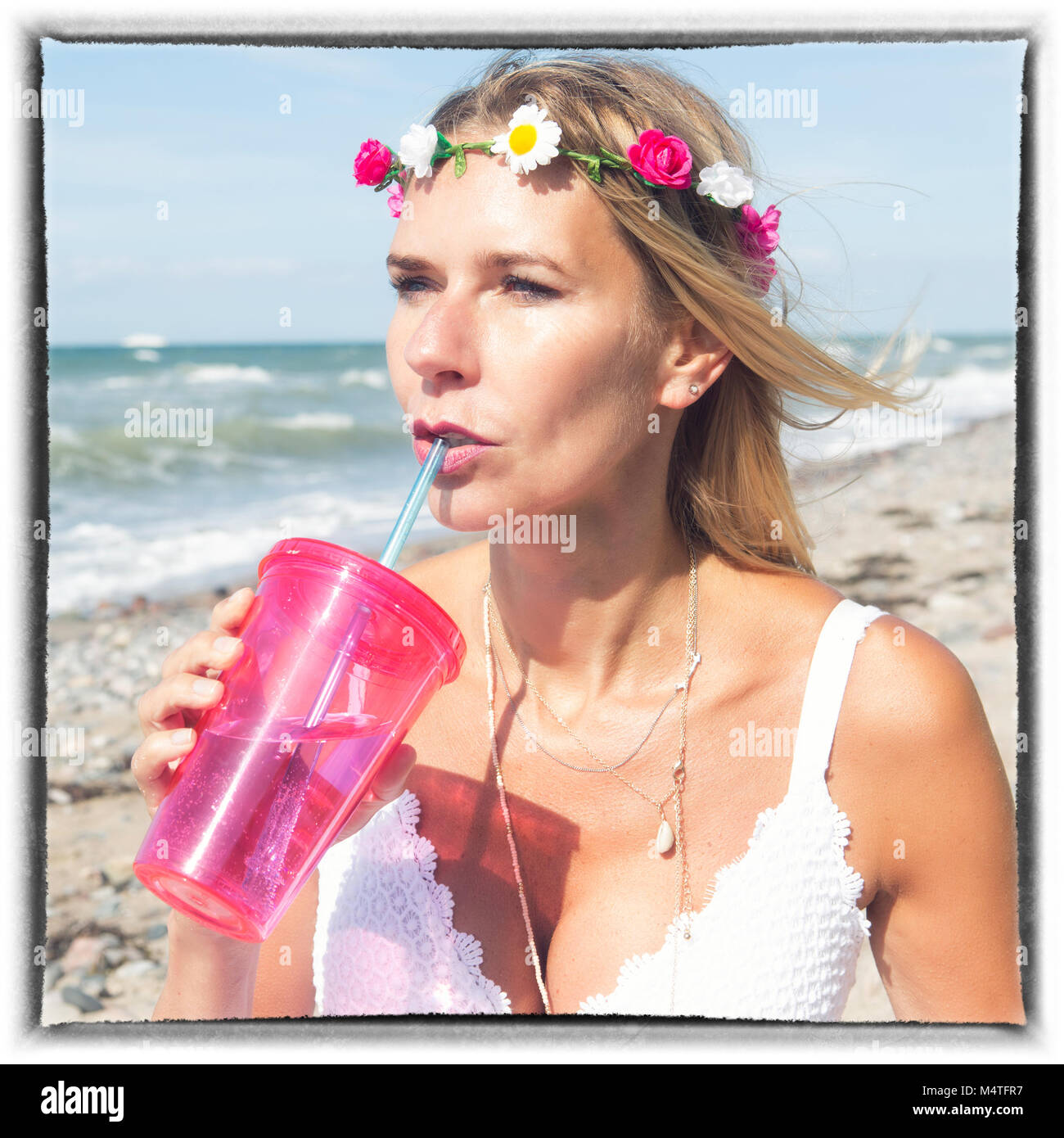 Donna bionda con fiori nei capelli e abito bianco permanente sulla spiaggia con bevanda Foto Stock