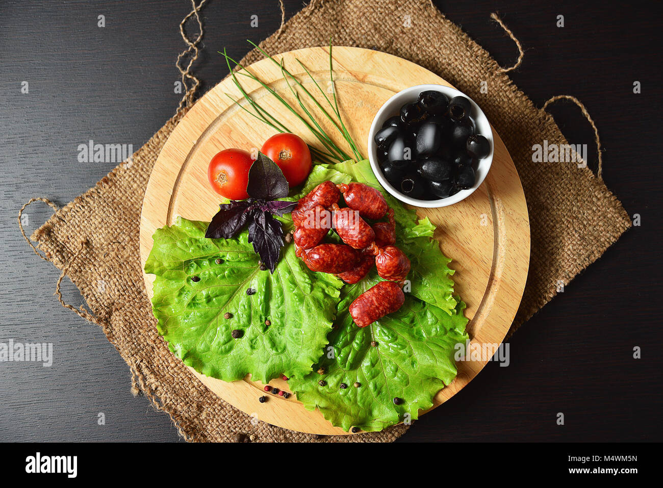 Salsicce di caccia su una tavola di legno con lattuga, basilico, pomodori ciliegini, paprika e olive. Foto Stock