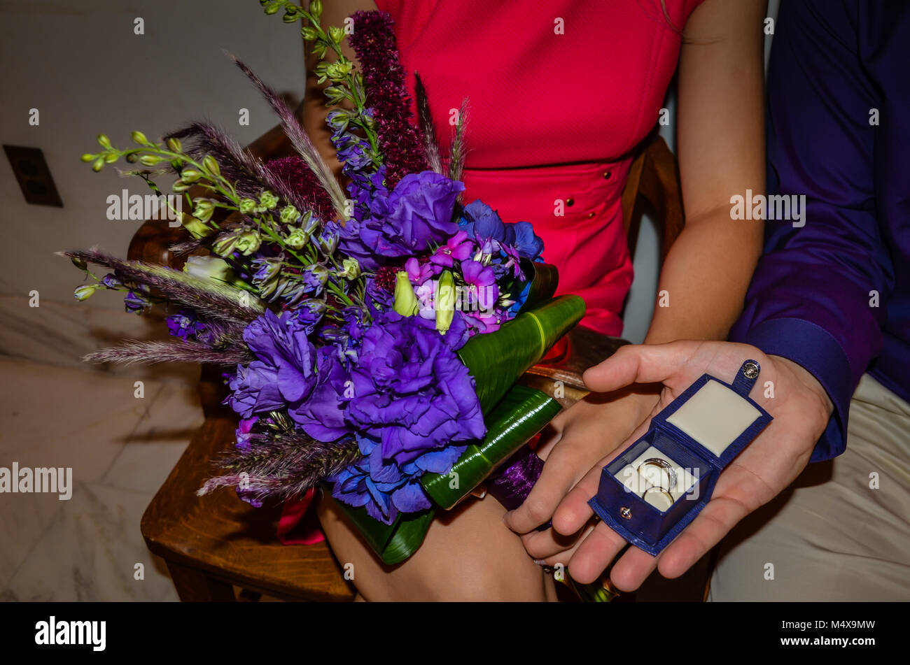 Gli anelli di nozze in un riquadro blu e viola bouquet detenute da una sposa in una rosa caldo vestito. Foto Stock