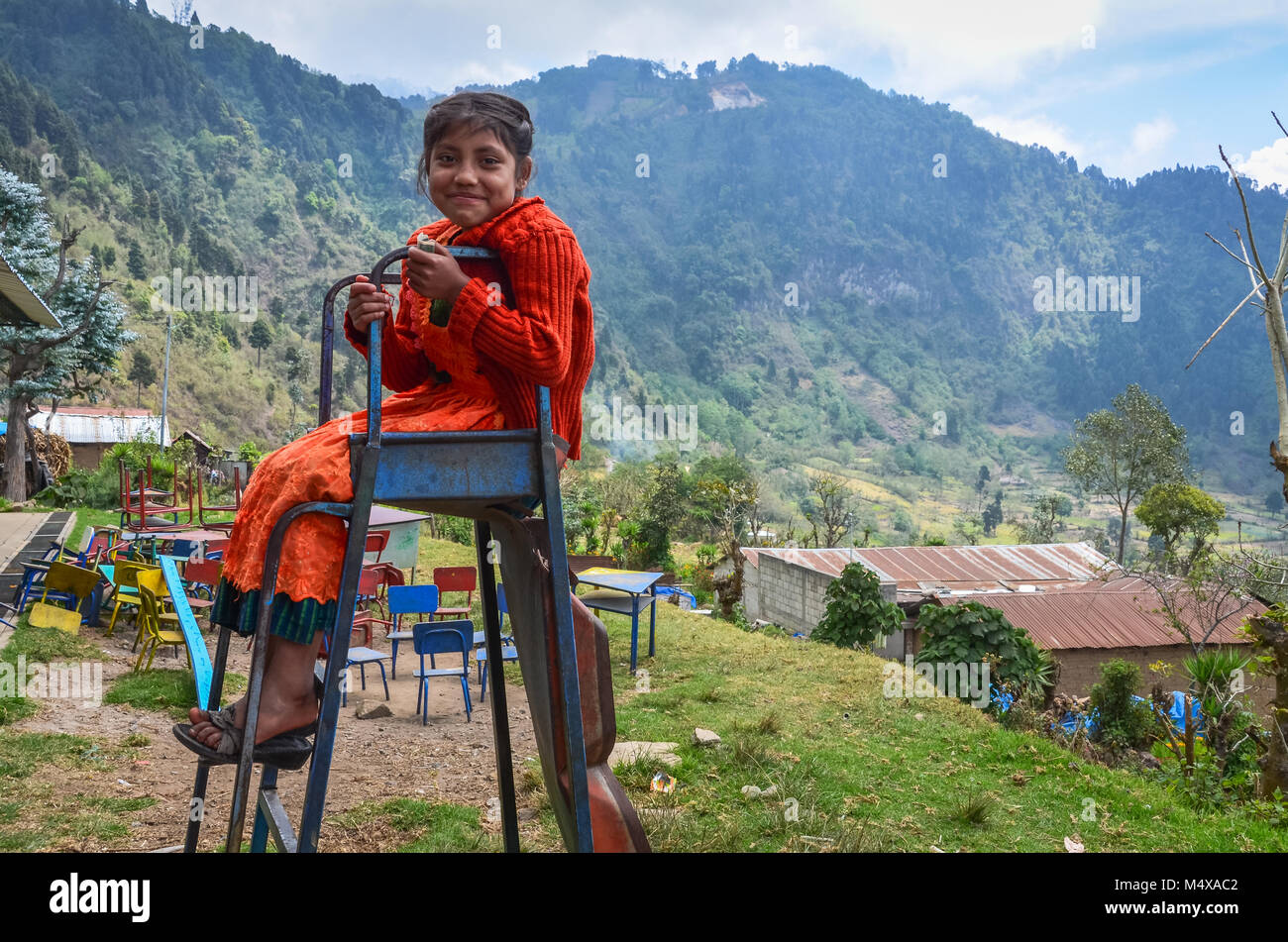 Sorridente giovane studente si siede in cima a una rotta in metallo scorrevole in corrispondenza di una zona rurale scuola guatemalteca dove tutti gli abitanti del villaggio vivono in povertà. Foto Stock