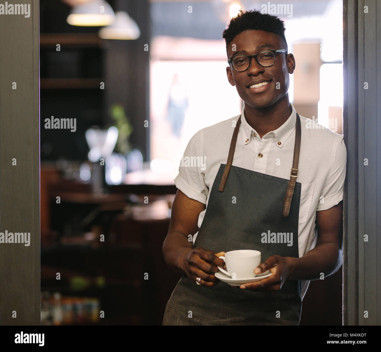 Vista dettagliata del barista in piedi all'ingresso del coffee shop tenendo una tazza di caffè. Caffè felice proprietario di un negozio di posa con una tazza di caffè in mano. Foto Stock