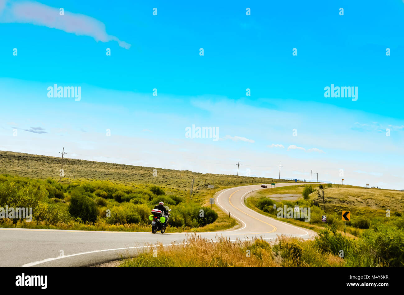 Un motociclista scorre giù per una strada tortuosa che le curve in avanti e verso l'alto, niente ma praterie su entrambi i lati e un grande cielo blu sopra. Foto Stock